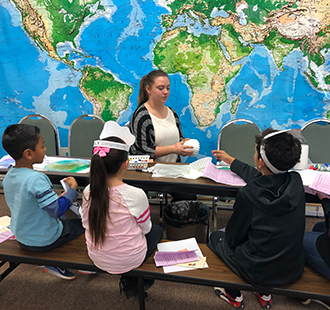 Teacher teaches children in class in front of a world map on a wall